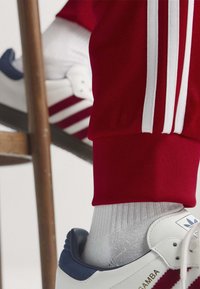 Λευκό αθλητικό παπούτσι Adidas Samba με κόκκινες ρίγες, φορεμένο με λευκές κάλτσες και κόκκινο παντελόνι με λευκές ρίγες, δίπλα σε ξύλινο πόδι καρέκλας.