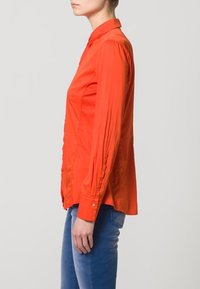 Camicia arancione con colletto a punta, maniche lunghe e vestibilità comoda, realizzata in un tessuto morbido. Indossata con jeans blu.