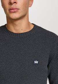 Ciemnoszary sweter z dzianiny z prążkowanym okrągłym dekoltem i małą okrągłą naszywką z logo na klatce piersiowej. Miękka tekstura i lekka konstrukcja.