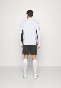 Îmbrăcăminte sportivă care include un top alb cu mânecă lungă și panouri laterale negre, pantaloni scurți închisi la culoare, șosete albe până la genunchi și încălțăminte sportivă cu accente neon.