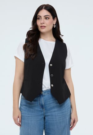 Kvinna med långt mörkt hår bär silverfärgade ringörhängen, en vit kortärmad topp, en svart knäppt väst och blå jeans med hög midja.