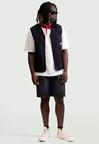 Gilet noir sans manches porté sur un t-shirt blanc, associé à un short en jean sombre et des baskets blanches. Accessoires : lunettes de soleil noires et bandana rouge.