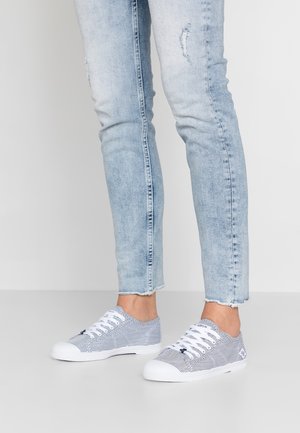 Benen dragen lichtblauwe gescheurde skinny jeans en lage witte sneakers met blauwe strepen en witte veters op een effen vloer.