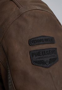 Giacca in pelle marrone con dettagli di texture. Presenta due toppe ricamate: "FLYING RELIC" e "PME LEGEND AMERICAN CLASSIC."