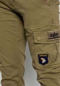 Olive green cargo pants made of cotton, featuring a textured fabric, side pockets, and emblems that include "Alpha Industries" and "Airborne."