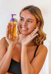Smiling woman in black bra holding a bottle of "glow your own way" hydrating body and face lotion with purple pump dispenser.