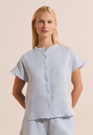 Blonde vrouw draagt een lichtblauw gestructureerd kort mouwtjes knoopoverhemd en bijpassende korte broek tegen een beige achtergrond.