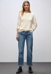 Cremfarbener, flauschiger Strickpullover mit langen Ärmeln, kombiniert mit hellblauen, geraden Jeans und schwarzen Schnürschuhen.