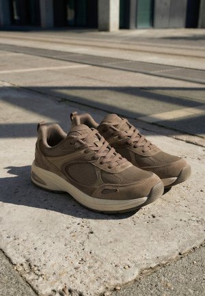 Brune snørestøvler med mesh- og ruskindsdetaljer, der hviler på betonbelægning under sollys.