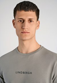Jasnoszara bawełniana koszulka z okrągłym dekoltem i czarnym napisem "LINDBERGH" na piersi. Gładka faktura i dopasowany krój.