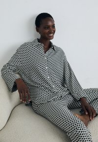 Smiling person with short hair seated on light sofa, wearing matching black and white geometric patterned shirt and pants.