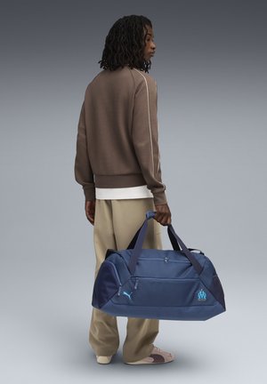 Sac de sport bleu marine en toile avec deux anses, poche zippée à l'avant, logo brodé et une texture lisse et durable. Idéal pour le sport ou les voyages.