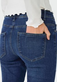 Tumed sinine denimist teksapüksid, millel on keha lähedane lõige, rõhutatud tagataskud ja peened õmbluse detailid. Kanga pind on sile.