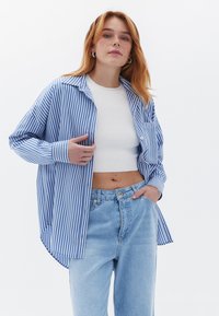 Gestreifte blaue und weiße Hemdbluse mit langen Ärmeln, lockerer Passform und abgerundetem Saum, getragen über einem weißen Crop-Top und hellblauen Jeans.