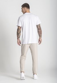 Biela bavlnená tričko, béžové špicaté nohavice a biele tenisky; obsahuje tetovania na rukách a neutrálny pozadie.