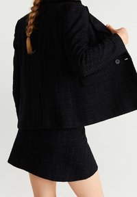 Veste noire texturée avec une jupe assortie cintrée. La veste présente une coupe structurée et des détails de boutons. Les deux pièces sont confectionnées dans un tissu tissé.