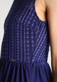Navyblaues Kleid mit strukturiertem Oberteil und geometrischen Mustern, ärmellosem Design und einem gerafften Rock. Inklusive eines kleinen Hardware-Akzents.