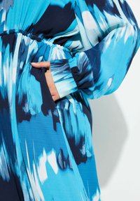 Une robe à motifs bleus et navy avec des manches longues, dotée d'une taille élastique et d'une poche latérale, fabriquée en tissu lisse à la texture fluide.