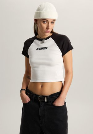 Młoda kobieta w białej czapce typu beanie i czarno-białym krótkim topie Vans, ubrana w czarne spodnie, ręce w kieszeniach, z przekłutym pępkiem.