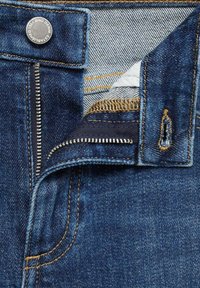 Primer plano de unos vaqueros vaqueros azules con cremallera plateada parcialmente abierta y botón de metal con la etiqueta "1991 MNG Denim Establisher."