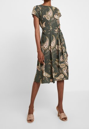 Vrouw draagt een groene midi-jurk met korte mouwen, paisley patroon en een bijpassende stoffen riem gestrikt in de taille, gecombineerd met beige sandalen met hak.