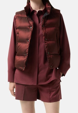 Woman wearing a burgundy button-up shirt, matching shorts, and a sleeveless dark red puffer vest with snap buttons, standing with one hand in pocket.