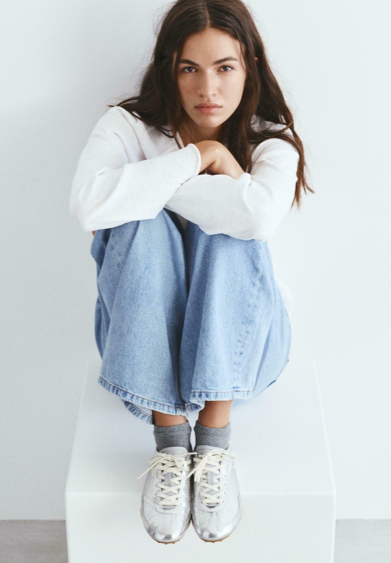 Fiatal nő hosszú sötét hajjal, fehér blokkban ülve, térdét ölelve, fehér pulóvert, világoskék farmert, szürke zoknit és fehér tornacipőt viselve.