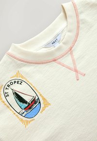 Krémově zbarvený žebrovaný svetr s růžovým prošitím, grafika plachetnice s nápisem "ST TROPEZ" a značková štítek "NEXT" uvnitř límce.