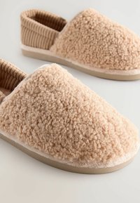 Beige Hausschuhe mit einem strukturierten, flauschigen Obermaterial und einem gerippten Knöchelbereich. Die Sohle ist hell gefärbt, flach und robust. Weiches, gemütliches Design.