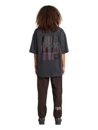 Osoba o kręconych włosach, ubrana w oversize'ową ciemnoszarą koszulkę z nadrukiem i ciemnobrązowe spodnie z numerem „223” na dolnej części nogawki, stoi tyłem.