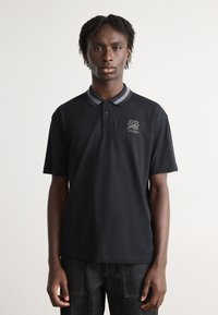 Zwarte polo met een grijs gestreepte kraag, voorzien van een geborduurd logo op de borst. Gemaakt van een gladde, ademende stof.