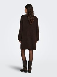 Oversized brown knitted dress with long sleeves, textured fabric, and a hem just above the knee; paired with tall black boots.