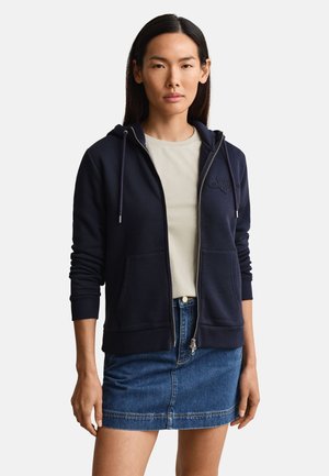 Žena odjevena u mornarsko plavu zip-hodu s džepovima sprijeda, bež košulju i plavu traper suknju, stoji ispred jednobojne pozadine.