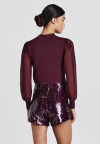 Top bordeaux con maniche a palloncino abbinato a pantaloni corti a vita alta in paillettes di una tonalità abbinata. Texture liscia con dettagli lucidi.