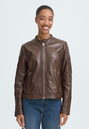 Smiling woman wearing a brown zipped leather jacket with two zip pockets and blue jeans, standing against a light background.