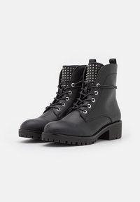 Botas negras hasta el tobillo con un upper de cuero texturizado, acentos de tachuelas plateadas en el cuello, cierre de cordones en la parte delantera y un tacón bloque grueso.
