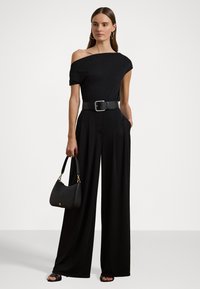 Black off-shoulder jumpsuit with wide leg design, cinched waist by a thick black belt, paired with a small black handbag.