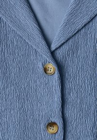 Veste en tissu bleu texturé avec un col cranté et deux boutons ronds marron fermés à l’avant.