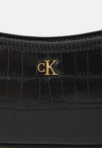 Zwarte nep-krokodillenleren handtas met een gebogen ontwerp. Beschikt over een gouden "ck" logo-accent en een gestructureerd oppervlak met horizontale stikseldetails.