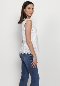 Top bianco senza maniche con spalline arricciate e dettagli in pizzo traforato all'orlo, abbinato a jeans in denim blu. Tessuto morbido.