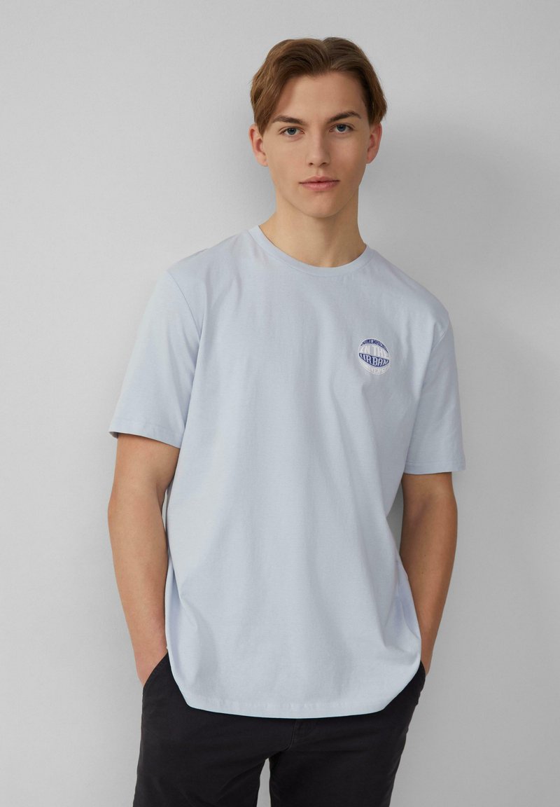 Lichtblauw katoenen t-shirt met een ronde halslijn, korte mouwen en een cirkelvormig logo-ontwerp op de linkerkant van de borst. Soepele textuur en een ontspannen pasvorm.