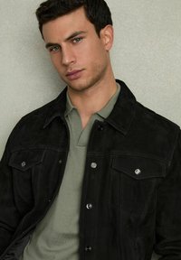 Jachetă din suede neagră cu butoane tip snap și buzunare, purtată peste un tricou polo de un verde deschis. Textură netedă, design casual, siluetă fitted.