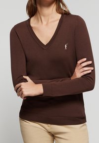Pull marron à col en V avec manches longues, poignets et ourlet côtelés, arborant un petit logo blanc sur le côté gauche de la poitrine. Tissu tricoté doux et texturé.