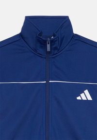 Mörkblå träningsjacka med hög krage, heldragkedja och vit horisontell randdekor. Har Adidas-logotypen på sidan.