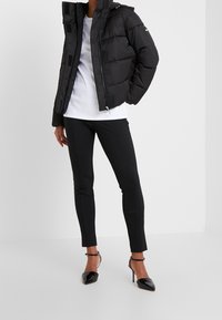 Giacca a vento nera con chiusura zip e cappuccio, abbinata a una camicia bianca, pantaloni neri a sigaretta e scarpe con tacco nere.