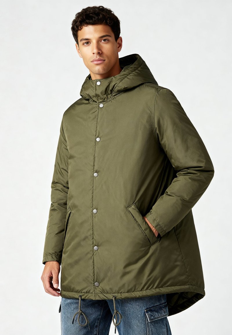 Homme portant un parka à capuche vert olive avec boutons-pression, poches latérales, et un jean cargo bleu, debout devant un fond uni.