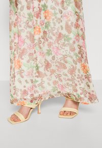 Vestido maxi floral con una tela ligera y translúcida que presenta patrones coloridos de paisley y flores. Combinado con sandalias de tacón alto desnudas y con tiras.