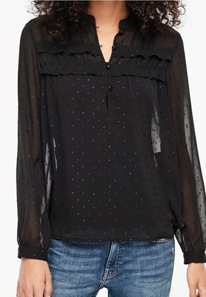 Blouse noire transparente à manches longues avec petit motif à pois, détails à volants sur la poitrine, col boutonné, portée avec un jean bleu.