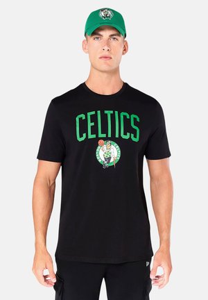 Uomo che indossa una t-shirt nera con testo e logo verdi "Celtics" e un cappellino verde con lo stesso logo, in piedi davanti a uno sfondo neutro.