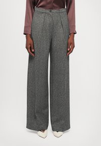 Donna che indossa pantaloni grigi in tweed a gamba larga e vita alta, con pieghe frontali e scarpe bianche a punta, in piedi di fronte a uno sfondo bianco.